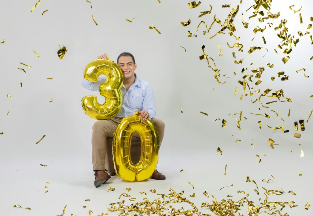 A man sitting on a chair holding a number three balloon surrounded by confetti