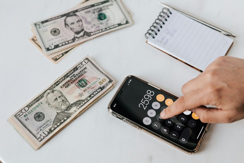 save money quickly on a tight budget -From above of crop anonymous economist calculating total amount of income using calculator app on cellphone near piles of greenbacks and notebook with pen