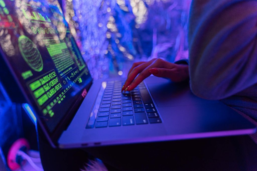 Spear Phishing- Close-up of hands typing on a laptop displaying cybersecurity graphics, illuminated by purple light.