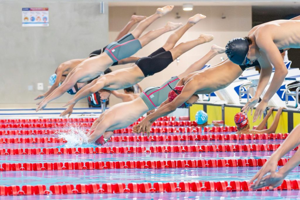 Swimmers dive into the pool during a competitive race in an indoor facility.