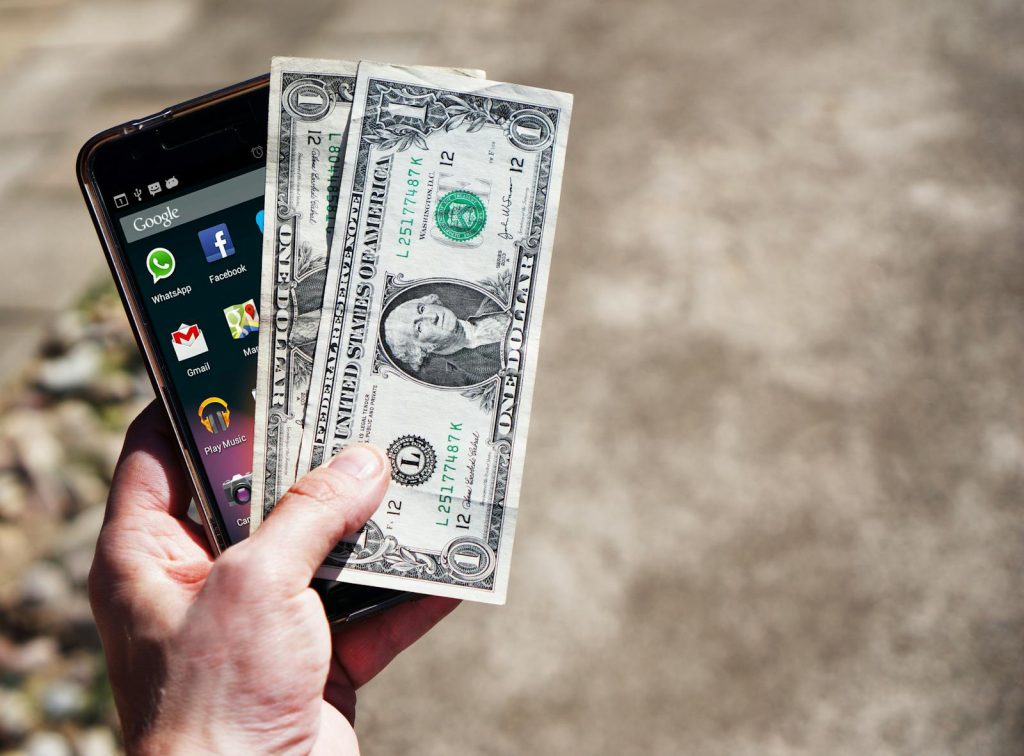 Government Cash Support- Close-up of a hand holding US dollar bills and a smartphone outdoors, showcasing financial technology.